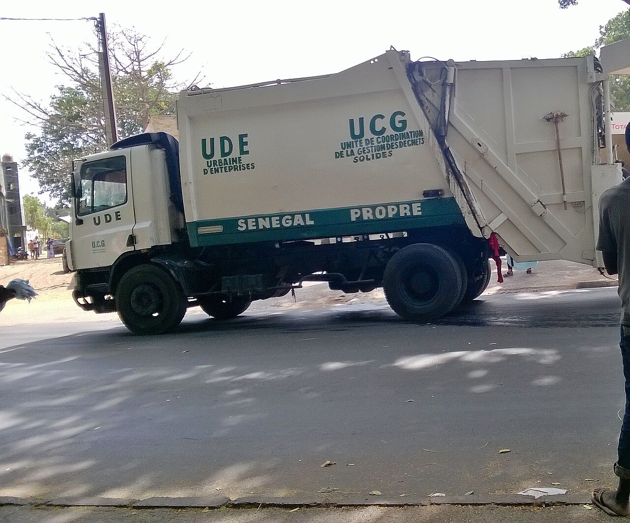 Un camion blanc de collecte des déchets est vu de côté sur une rue. Les inscriptions UCG, UDE et SÉNÉGAL PROPRE apparaissent sur la carrosserie ; des arbres bordent l’arrière-plan.