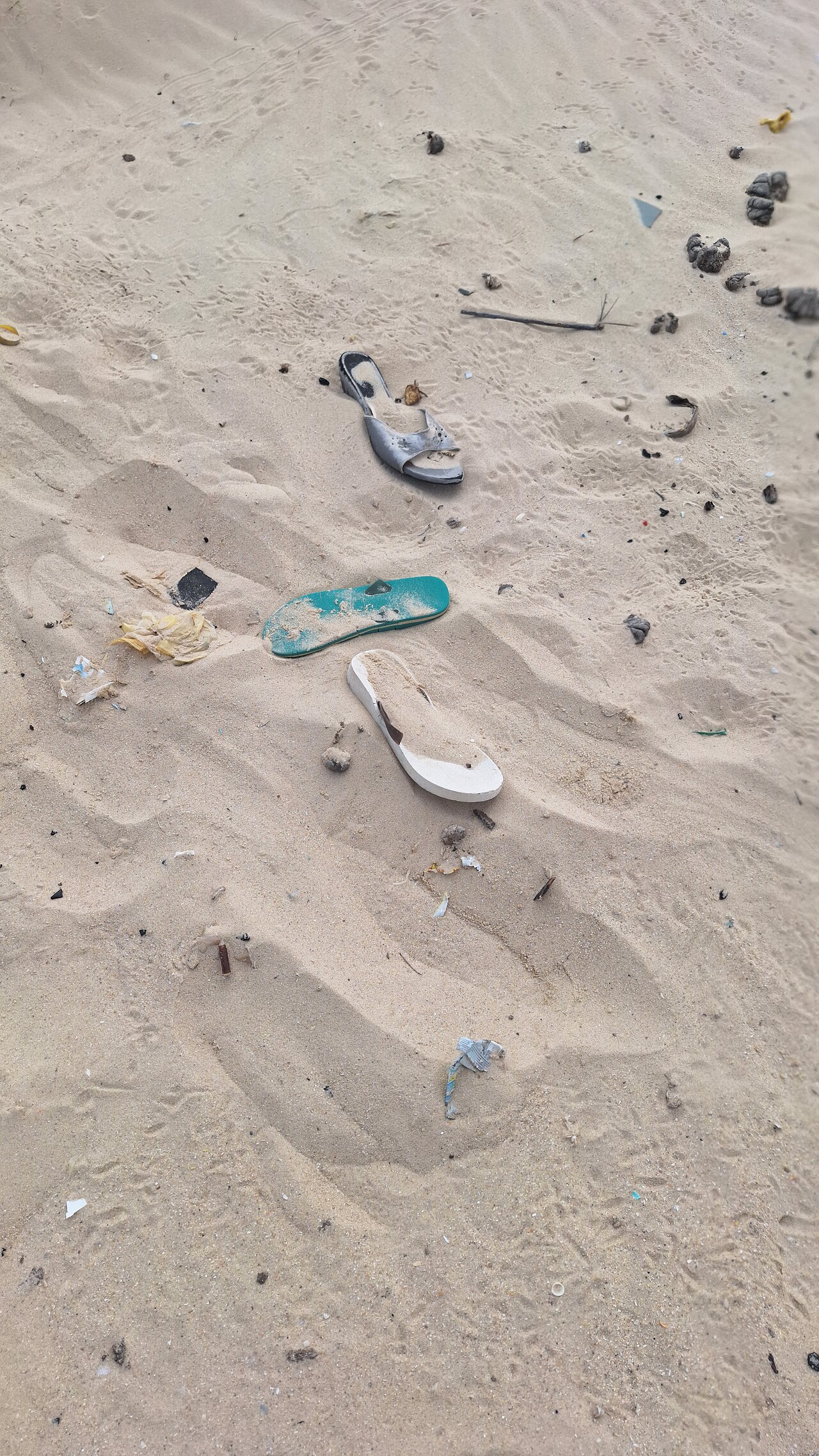 Sur du sable clair, plusieurs objets abandonnés sont visibles, dont une sandale turquoise et une chaussure ou semelle claire, avec de petits fragments dispersés.