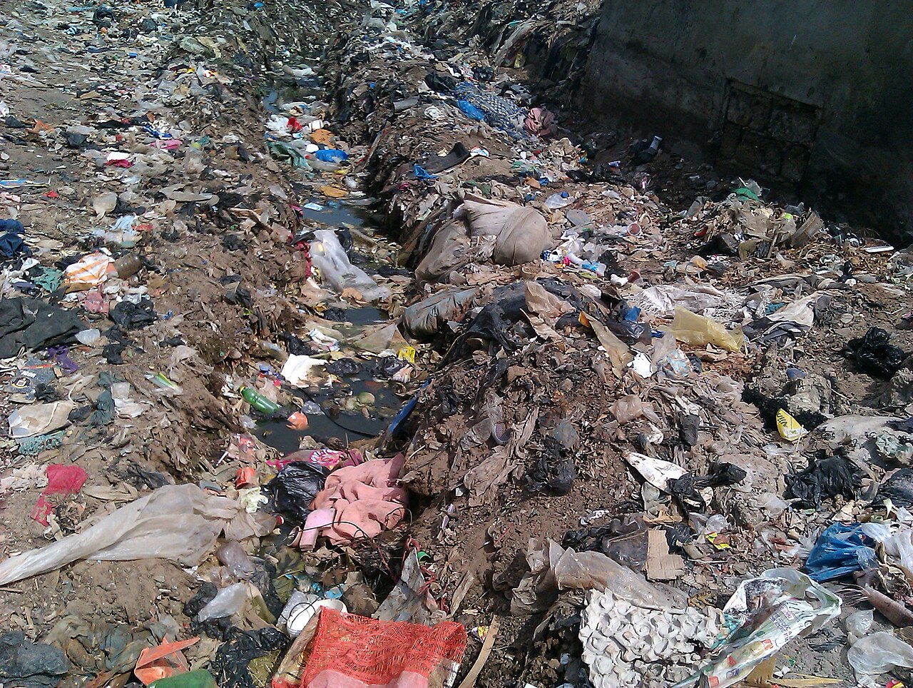Des déchets variés, des emballages colorés et des matières sombres jonchent un espace en creux au pied d’un mur en béton.
