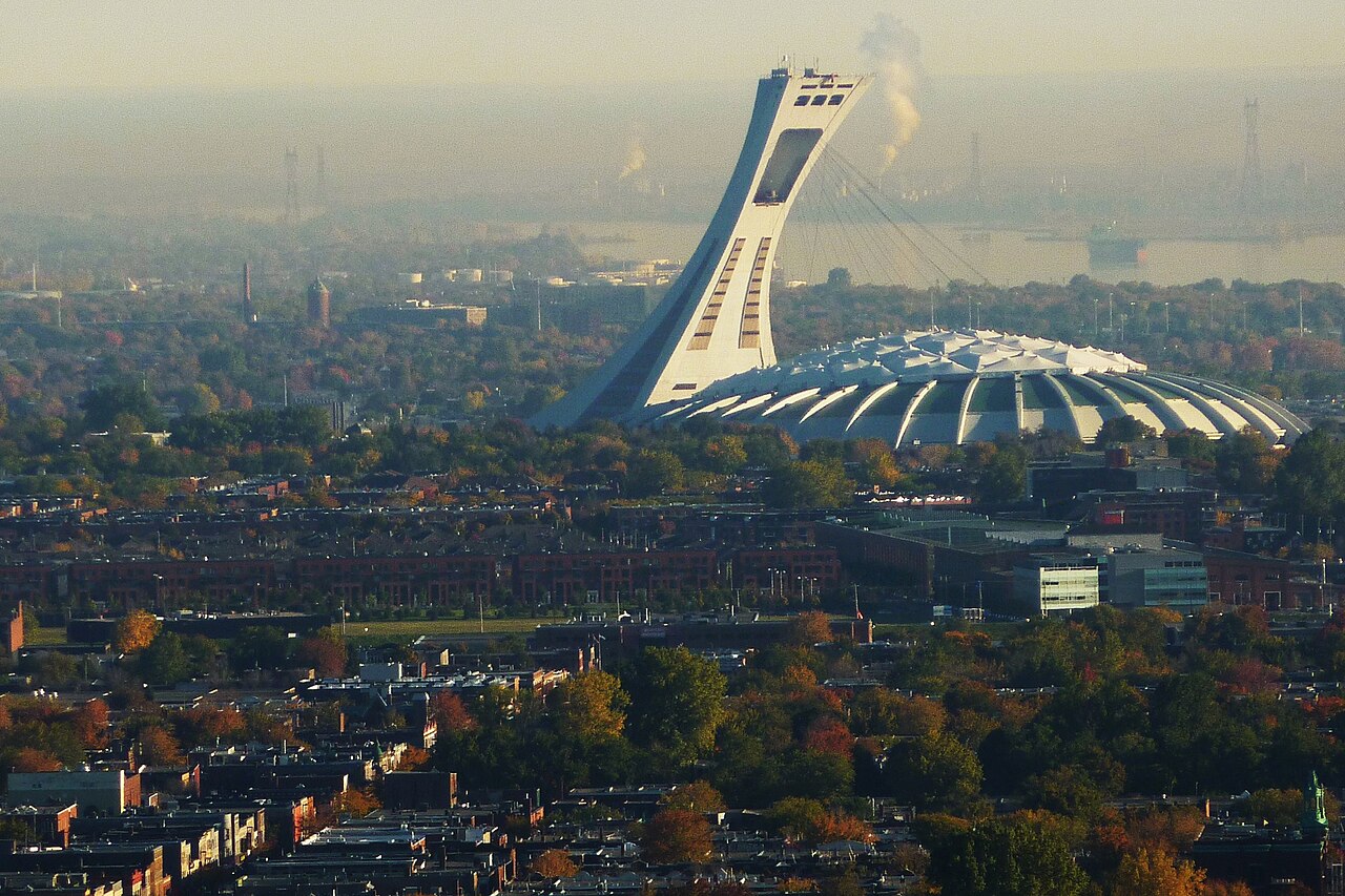 Une vue de Montréal montre le Stade olympique et sa grande tour inclinée au centre, des immeubles et des arbres au premier plan. Un voile trouble réduit la netteté des constructions lointaines.