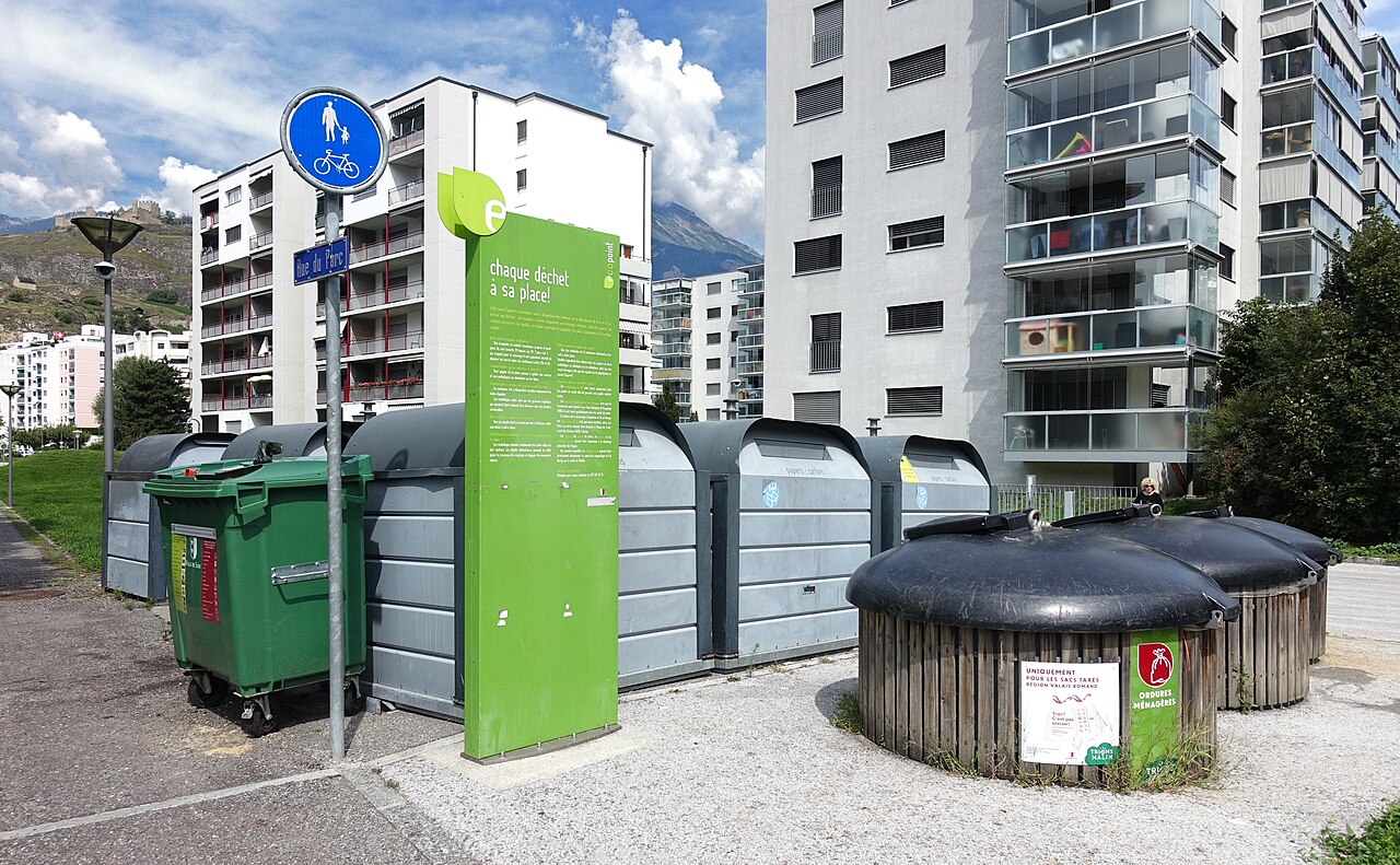 Un point de collecte regroupe des bacs verts, des conteneurs gris et des bornes rondes sombres. Un panneau vert se trouve au centre, devant des immeubles d’habitation et un relief montagneux au loin.