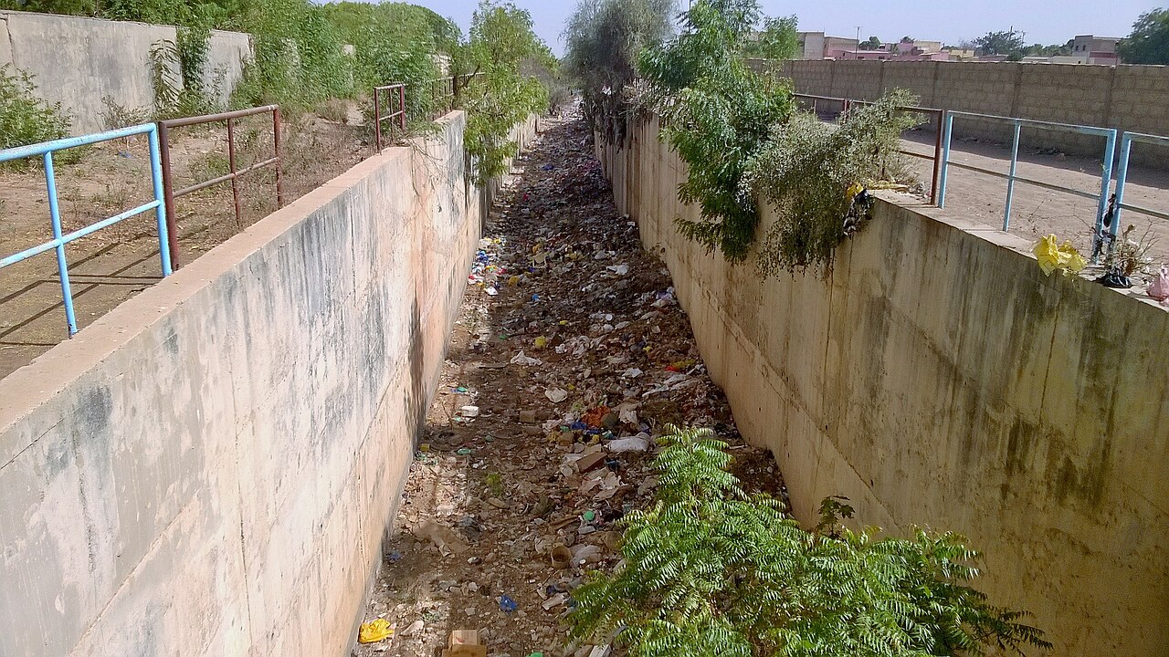 Un long canal de béton s’enfonce vers l’arrière-plan entre deux murs élevés. Des déchets et des fragments colorés couvrent le fond. Des garde-corps bleus et brun-rouille bordent les passages. De la végétation pousse au-dessus des murs et au fond, et des bâtiments sont visibles au loin.