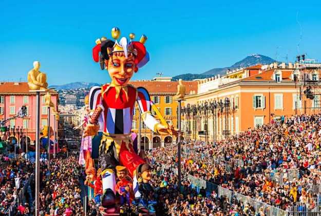 Une grande figure colorée domine le défilé de carnaval, avec des spectateurs de chaque côté et des façades aux tons chauds.